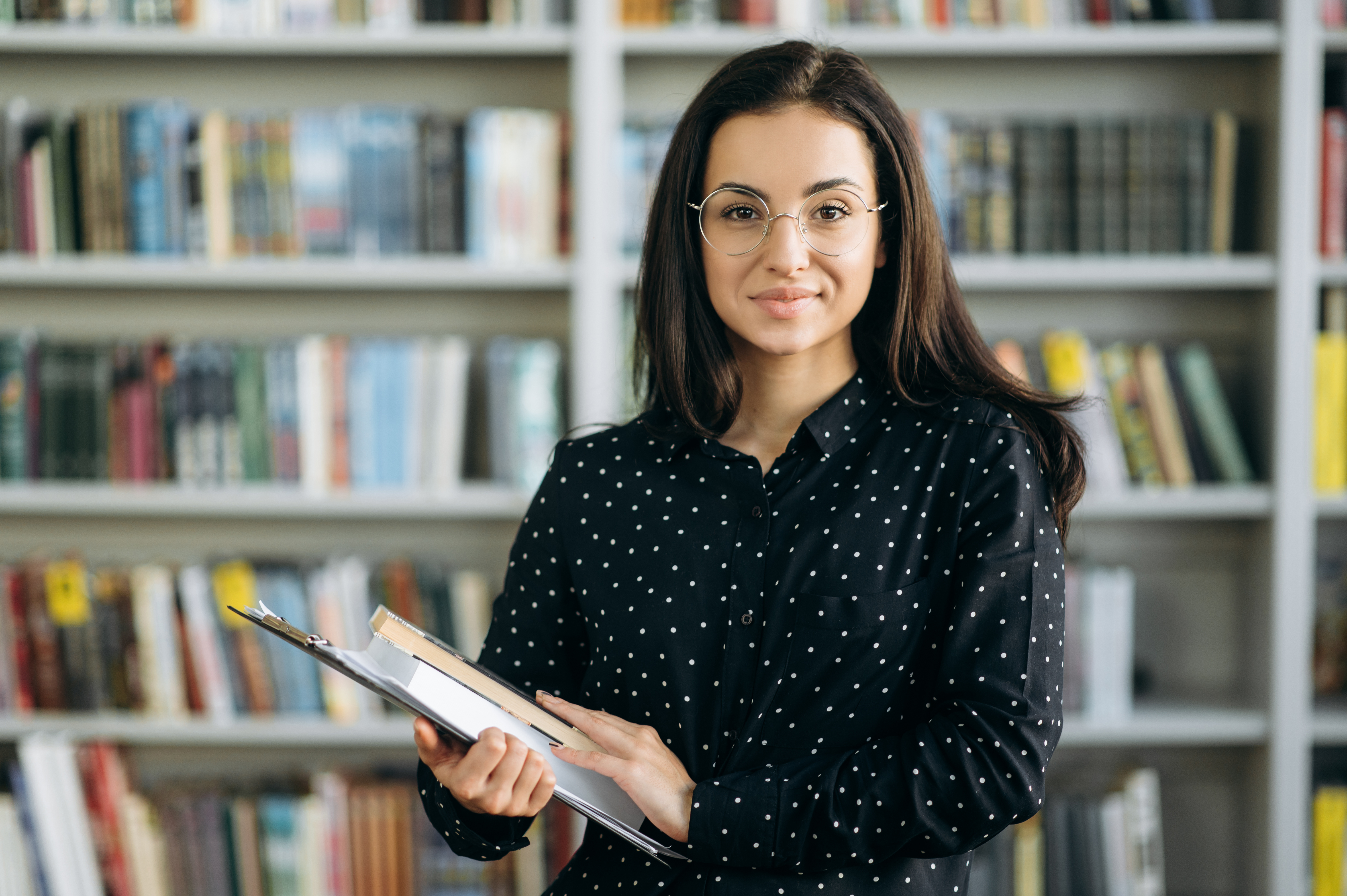 Portrait of an attractive confident young woman, student, teacher or business woman, in eyeglasses, looks directly at the camera with books in her hands, standing on books background