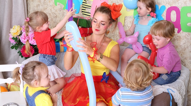 Five happy kids and facilitator make dogs of long balloons at children party. Inscription Happy Birthday on wall.